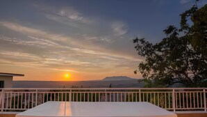 Ein Balkon mit einem weißen Tisch und Stühlen, der Blick geht auf einen Sonnenuntergang mit Bergen in der Ferne.