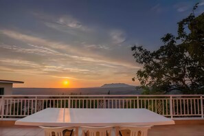 A balcony with a white table and chairs, overlooking a sunset with mountains in the distance.