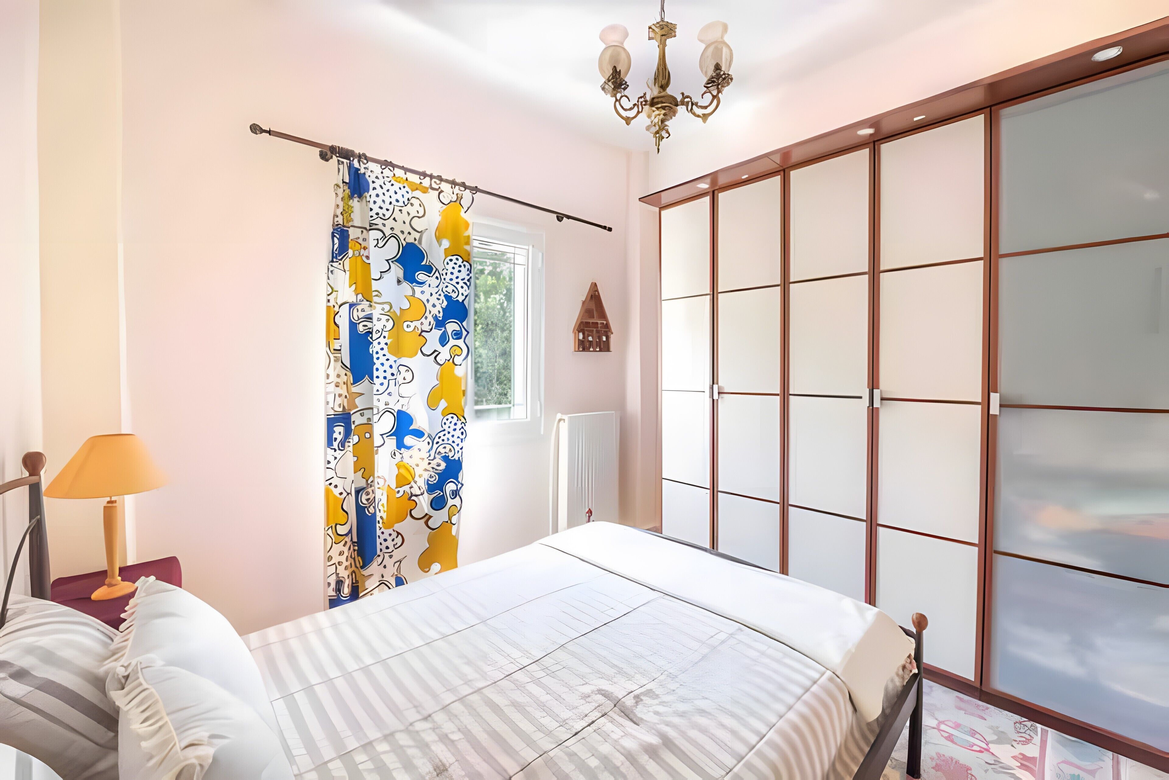 A bedroom with a bed, a window with a curtain, a radiator, and a chandelier.