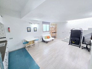 A spacious room with a white ceiling, a dining table with chairs, a white sofa with cushions, a wooden shelving unit, and a black office chair. - Oasis House (Maspalomas)