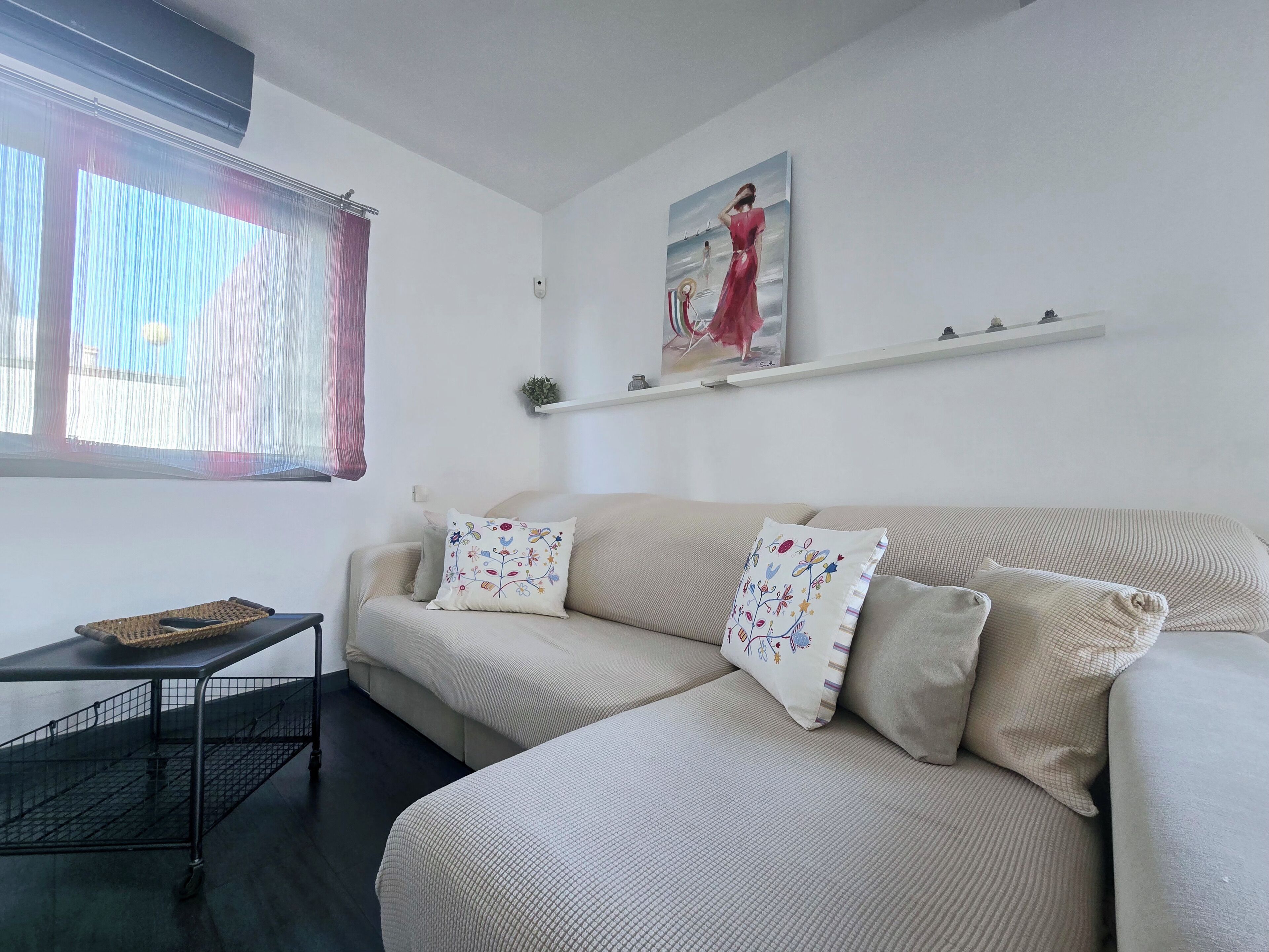 A living room with a sofa, two decorative pillows, a small table with a basket, and a painting on the wall.