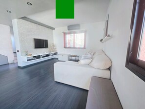A modern living room with a white sofa, a flat-screen TV mounted on a textured wall, a small white cabinet, and a window with curtains. - Oasis House (Maspalomas)