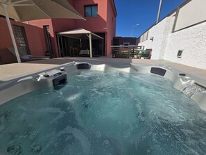 Ein Whirlpool mit Bedienfeld, umgeben von einer Terrassenanlage mit überdachter Sitzgruppe und einem Gebäude im Hintergrund.