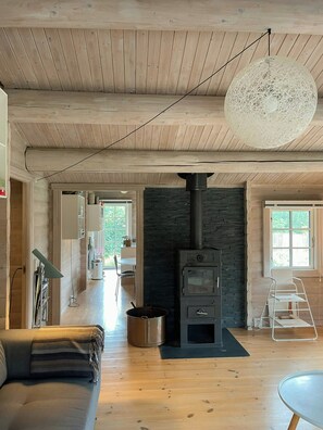 A cozy living room with a wooden ceiling, a stone fireplace, a sofa, and a dining area visible through an open door. - Beamed house by the water in Hornbæk (Hornbæk)
