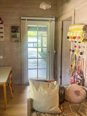 A room with a wooden floor, a table, a chair, a window with a view of a wooden deck, and a door with a towel rack holding colorful towels. - Beamed house by the water in Hornbæk (Hornbæk)