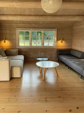 A cozy room with wooden floors, a sofa, a couch, two small tables, and a large window with a view of greenery. - Beamed house by the water in Hornbæk (Hornbæk)