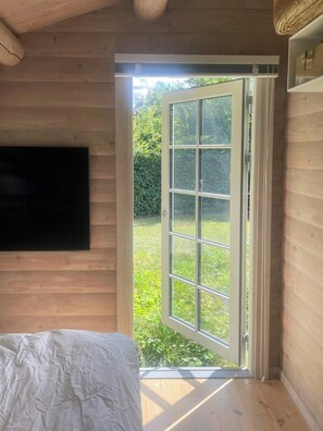 Quarto com paredes de madeira, uma cama, televisão montada na parede e uma porta de vidro deslizante que dá para um jardim.