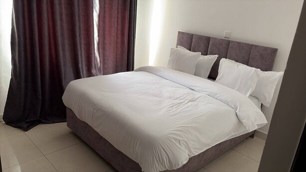 A bed with white bedding, a headboard, and a window with curtains. - Divine Homes Cosy 2 Bedroom Next to Airport Apartments (Nairobi)
