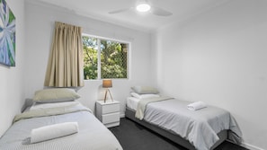 Ein Schlafzimmer mit zwei Einzelbetten, einem Nachttisch mit Lampe, einem Fenster mit Vorhängen und einem Deckenventilator.
