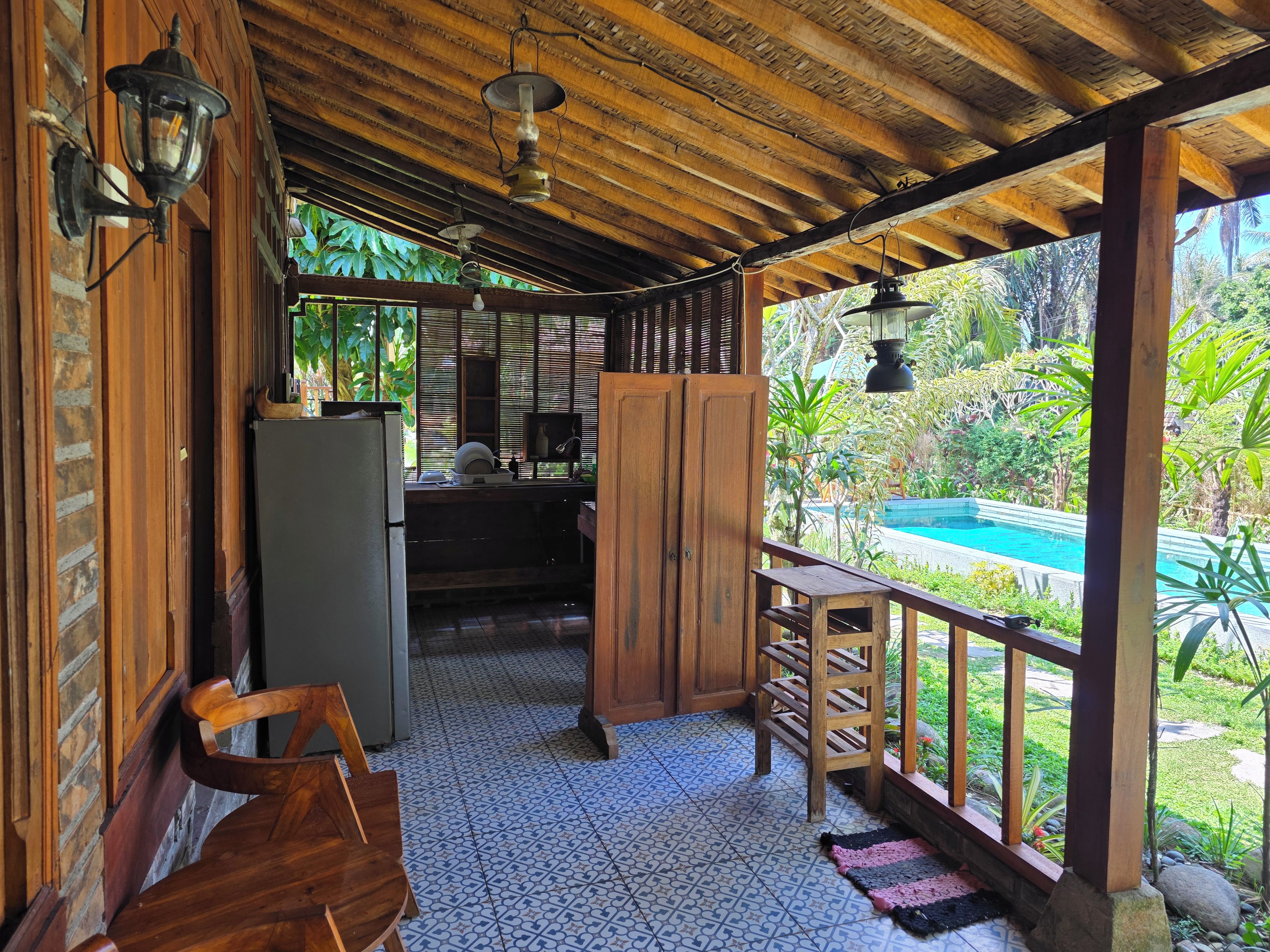 A covered outdoor area with a pool, wooden furniture, and a refrigerator.