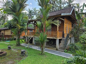A wooden bungalow with a balcony, surrounded by lush greenery and palm trees.
