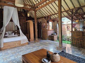 A room with a bed under a canopy, a wooden table with a vase and remote, and a brick wall with a door. - Sangeh Lodge (Abiansemal)