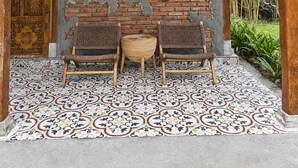A covered porch with two wicker chairs and a small table, a wooden door with intricate carvings, and a tiled floor with a geometric pattern.