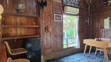 Una habitación de madera con una mesa de madera, dos sillas de mimbre, un tapete estampado, una puerta con vistas al exterior y un aire acondicionado fijado a la pared.
