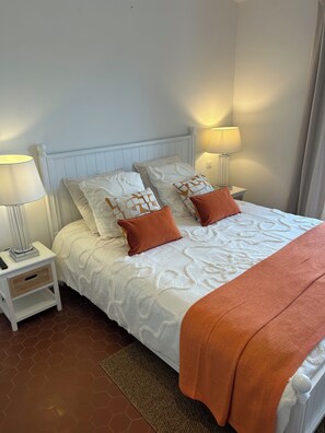 A neatly made bed with white bedding and orange pillows, flanked by bedside tables with lamps, in a room with a white headboard and plain walls. - A Beautiful House in Les Restanques (Grimaud)