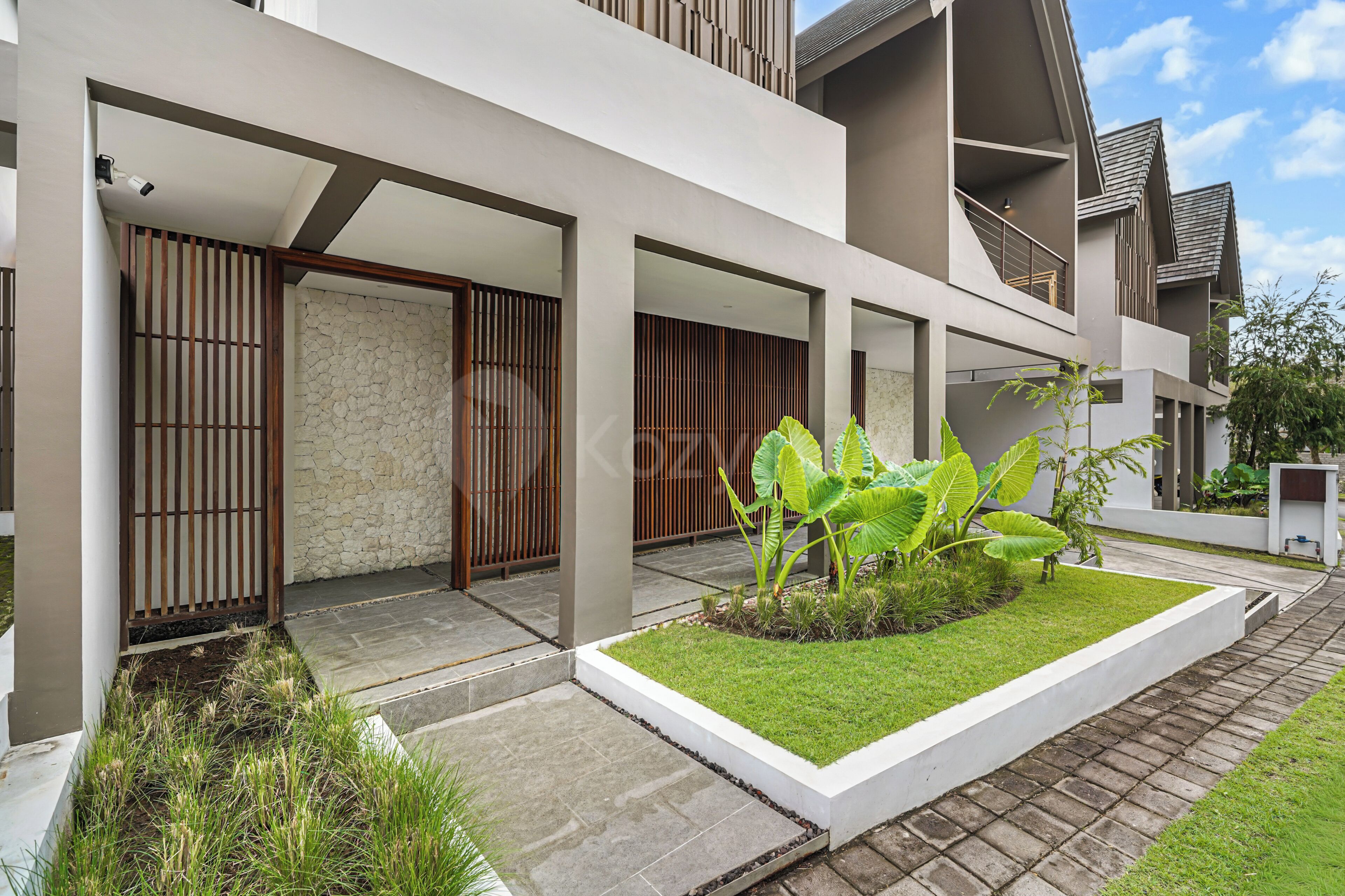 A modern building with a covered outdoor area, stone walls, and a small garden with tropical plants.
