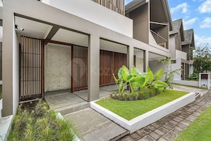 A modern building with a covered outdoor area, stone walls, and a small garden with tropical plants.