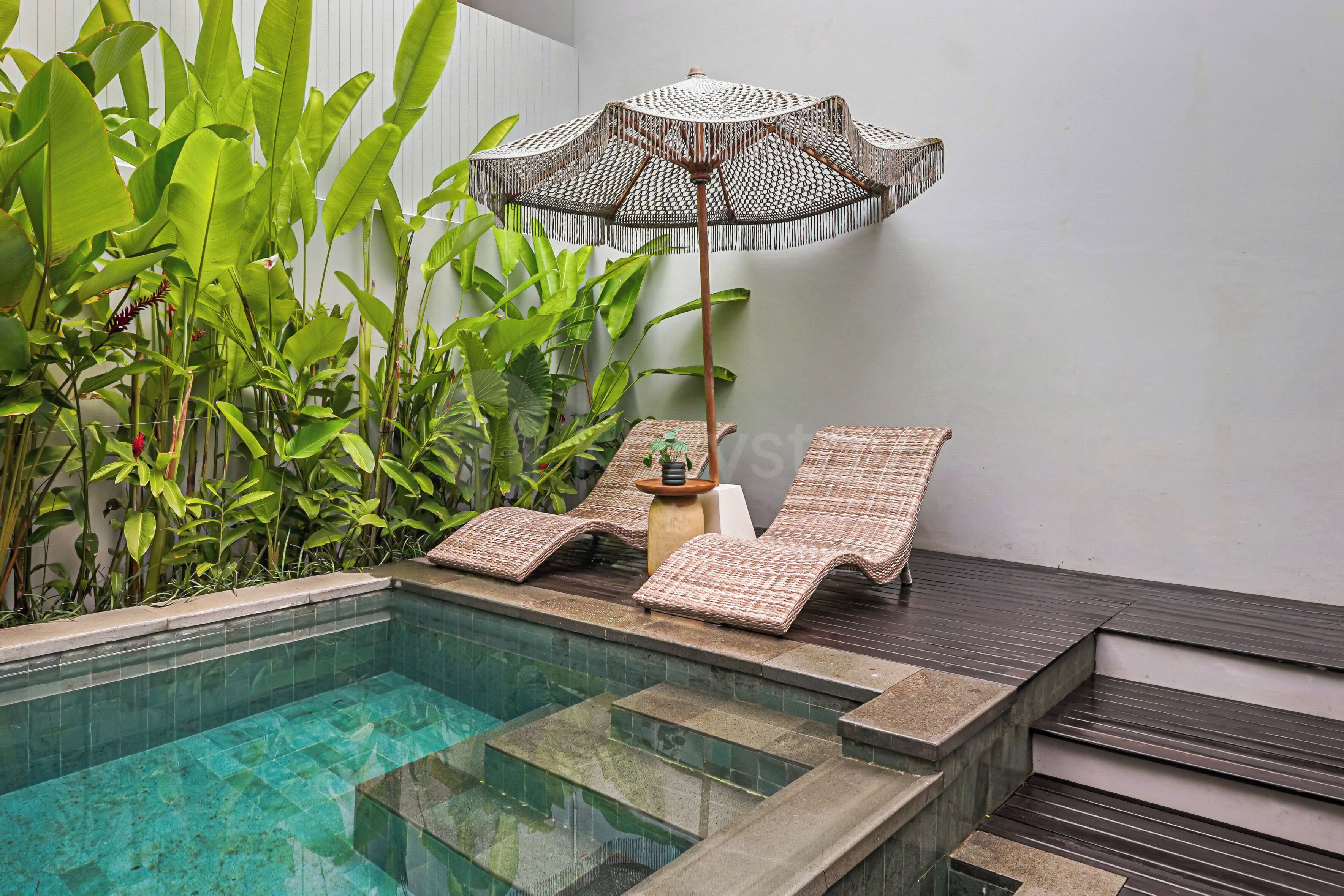 A pool area with a lounge chair and an umbrella, surrounded by lush green plants.