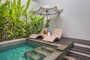 A pool area with a lounge chair and an umbrella, surrounded by lush green plants.