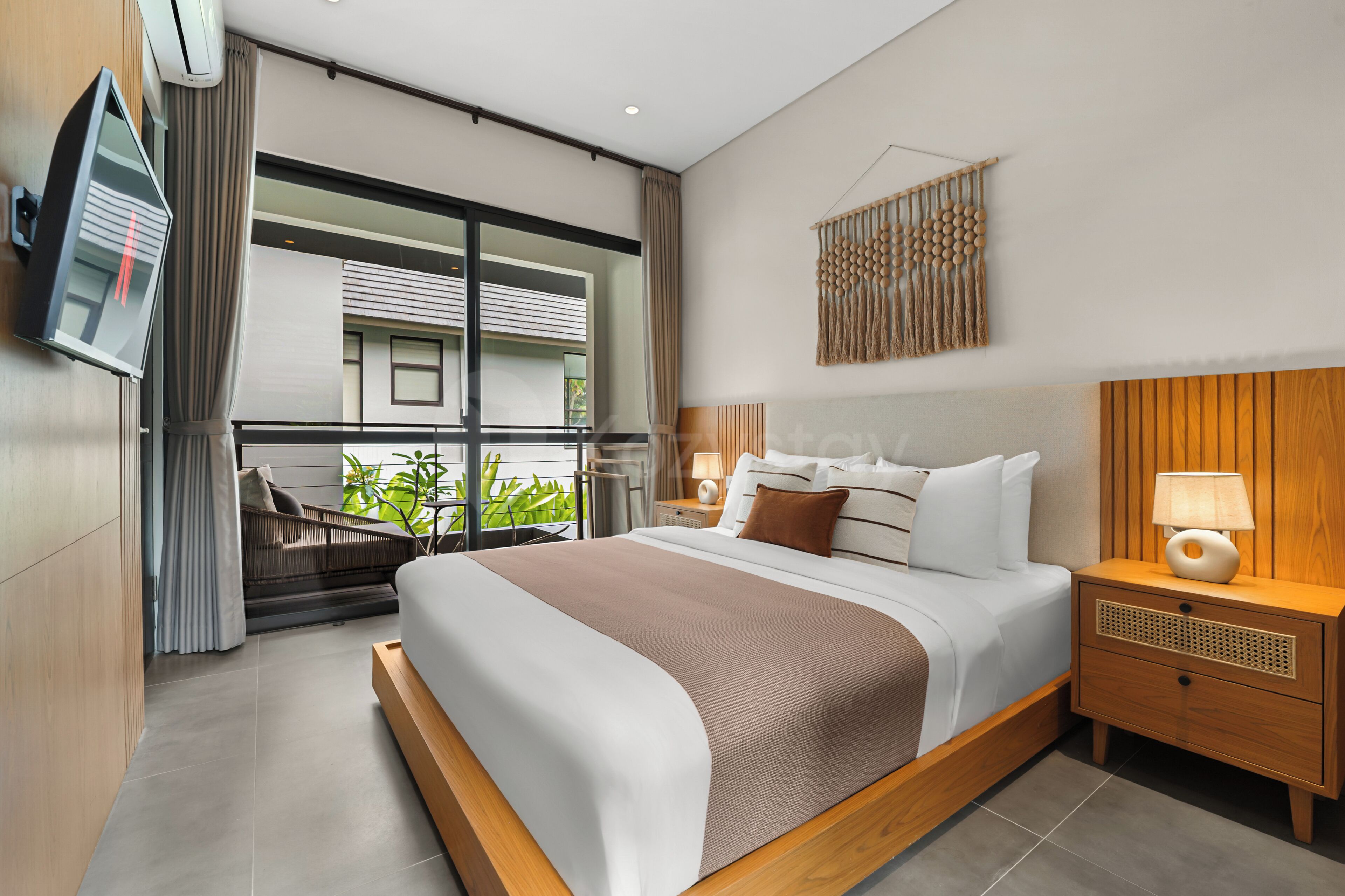 A modern hotel room with a large bed, a bedside table with a lamp, a TV mounted on the wall, and a view of the outside balcony.