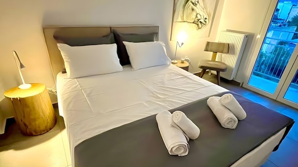 A hotel room with a large bed, a nightstand, a lamp, and a view of the city through a large window. - Kalliopi's Apartment with Stunning Acropolis View (Athens)