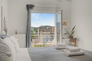 A bedroom with a bed, a radiator, a window with a view of buildings, a chair, and a plant. - Kalliopi's Apartment with Stunning Acropolis View (Athens)