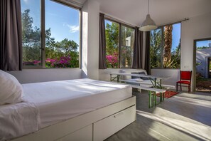 Ein modernes Schlafzimmer mit Bett, Tisch und Stuhl. Große Fenster bieten Blick auf Bäume und Blumen.