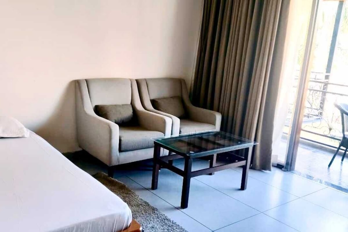 A hotel room with a bed, a sofa, a glass coffee table, and a view of the outdoors through a window with curtains.
