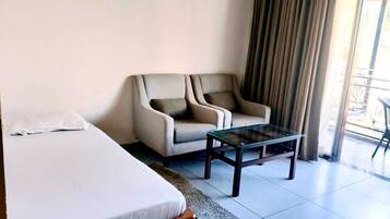 Habitación de hotel con cama, sofá, una mesita de centro de vidrio y vistas al exterior a través de una ventana con cortinas.