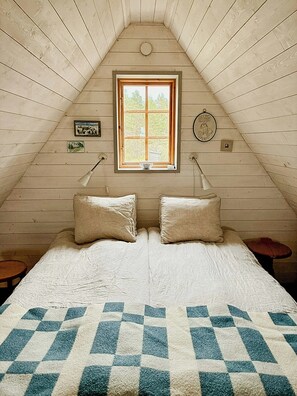 A room with a sloped ceiling, a window, two bedside tables, a bed with a checkered blanket, and wall-mounted lamps. - Traditional Gotland log houses (Gotlands län)