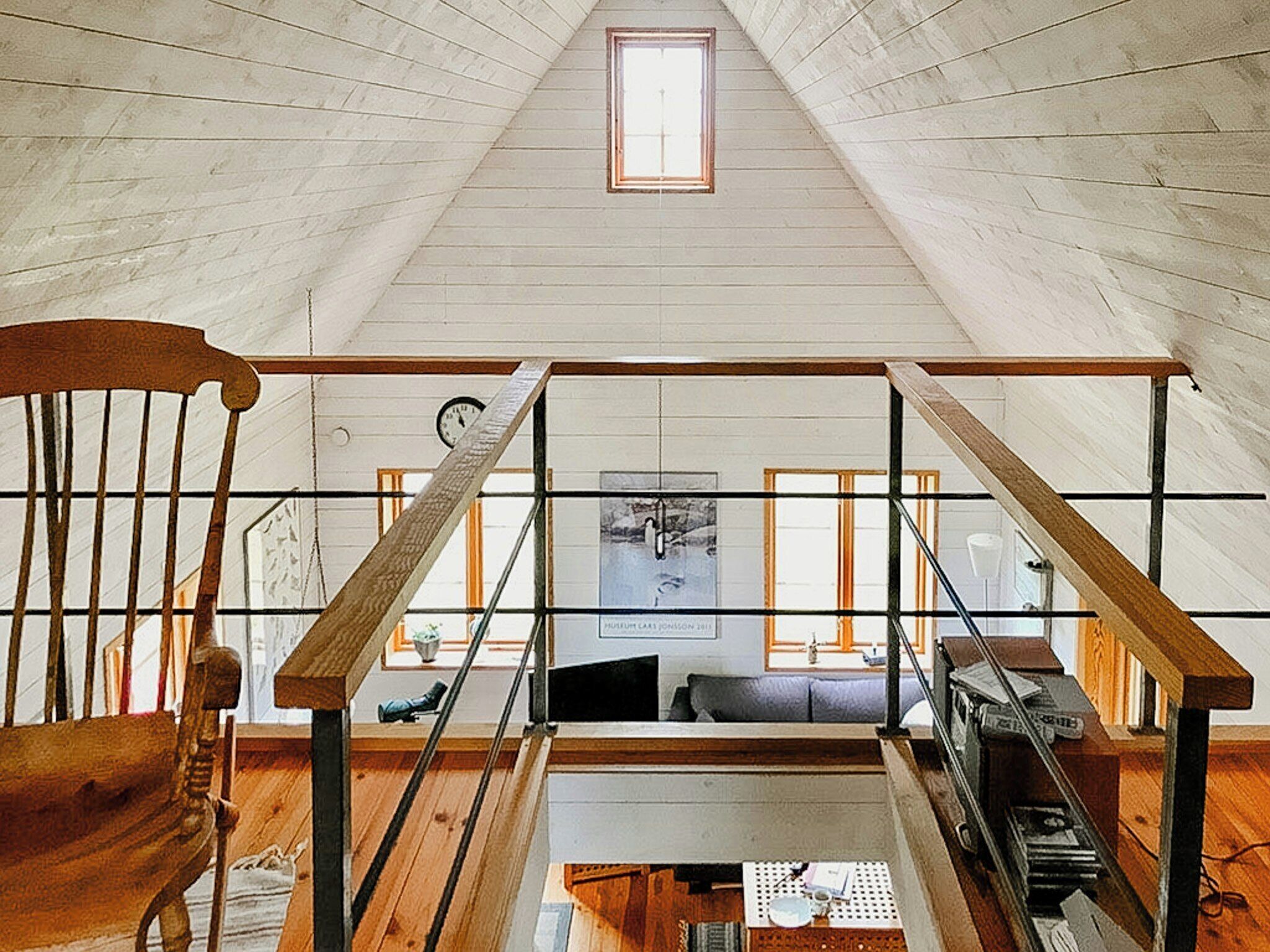 A loft bedroom with a sloped ceiling, wooden beams, a small skylight, a wooden chair, a bed with a headboard, and a window with a view of the outside.