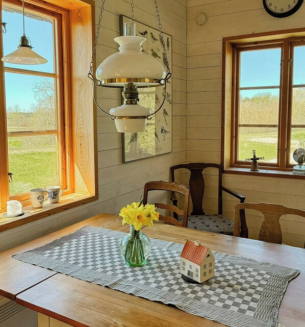 Een eetgedeelte met een tafel gedekt voor twee personen, een vaas met bloemen en een klein modelhuisje op een geruit tafelonderzetter.