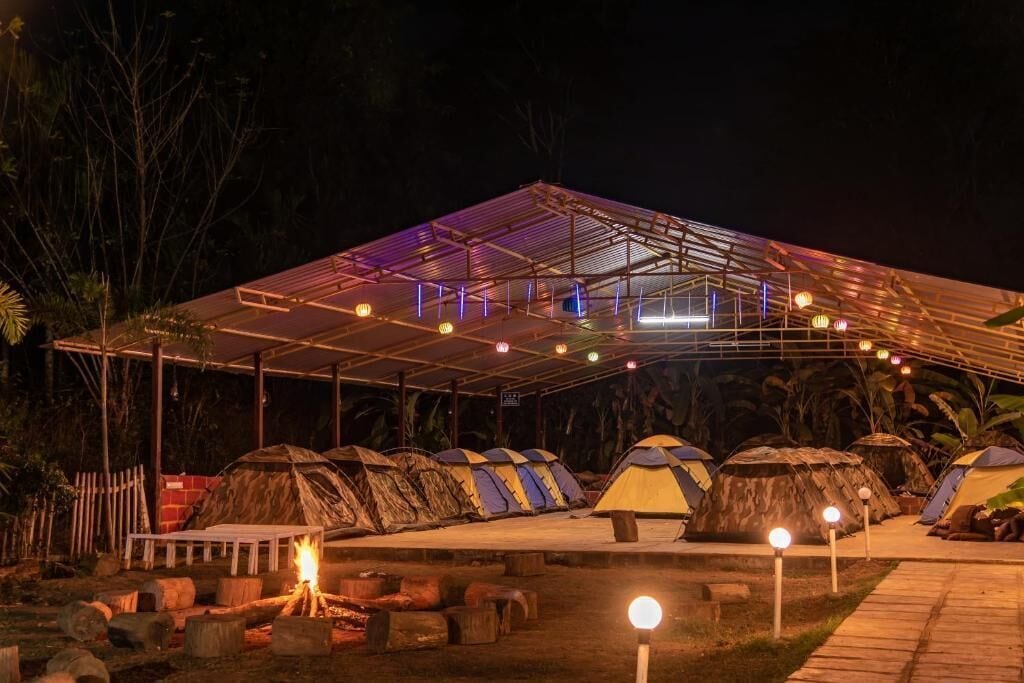 A covered outdoor area with multiple thatched-roof tents, a central fire pit, and a metal roof structure.