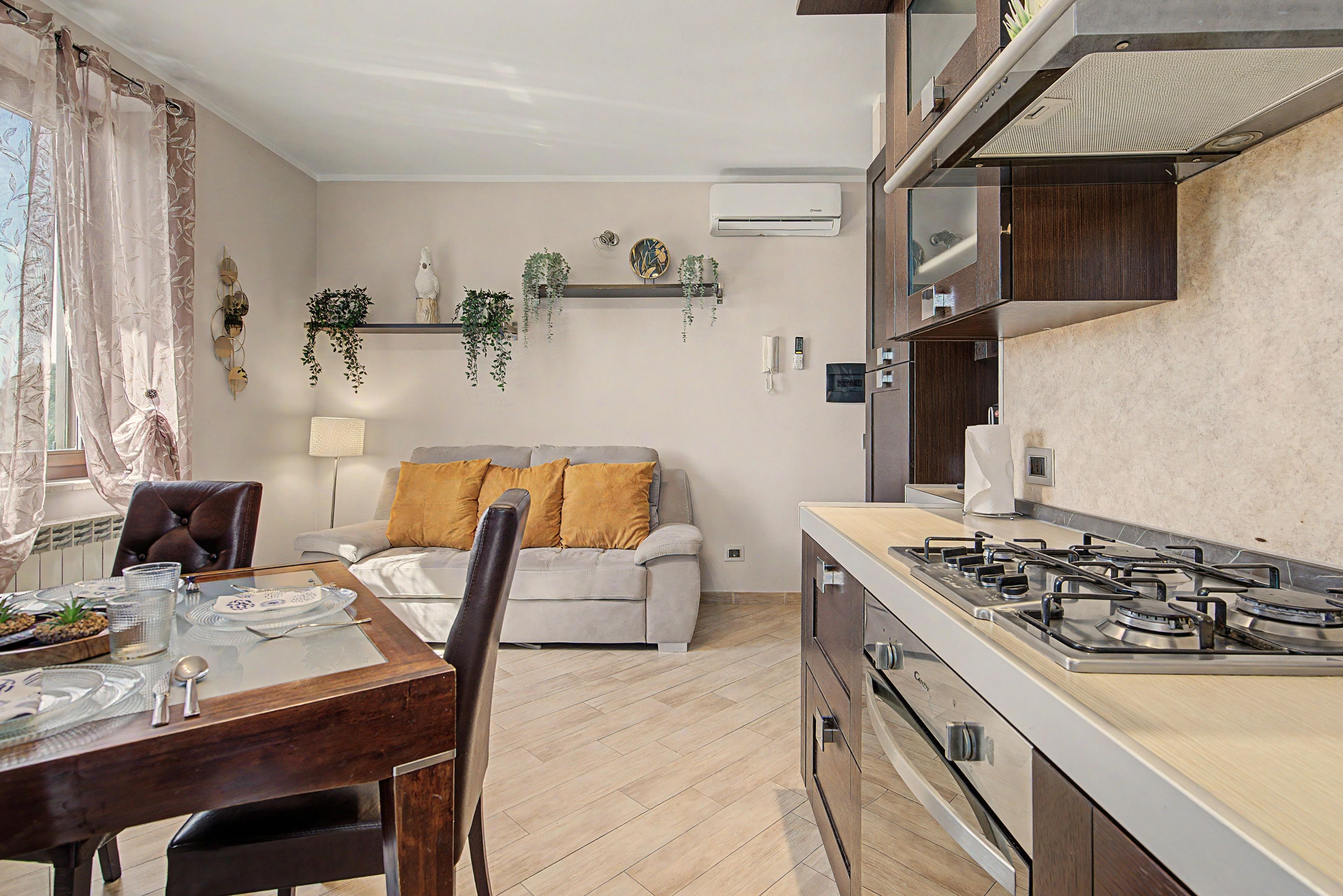 A modern kitchen with a gas stove, a dining area with a table set for two, and a living area with a sofa and cushions.