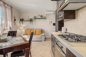 Una cuina moderna amb fogó a gas, zona de dinar amb taula per a dues persones, i saló amb sofà i coixins.