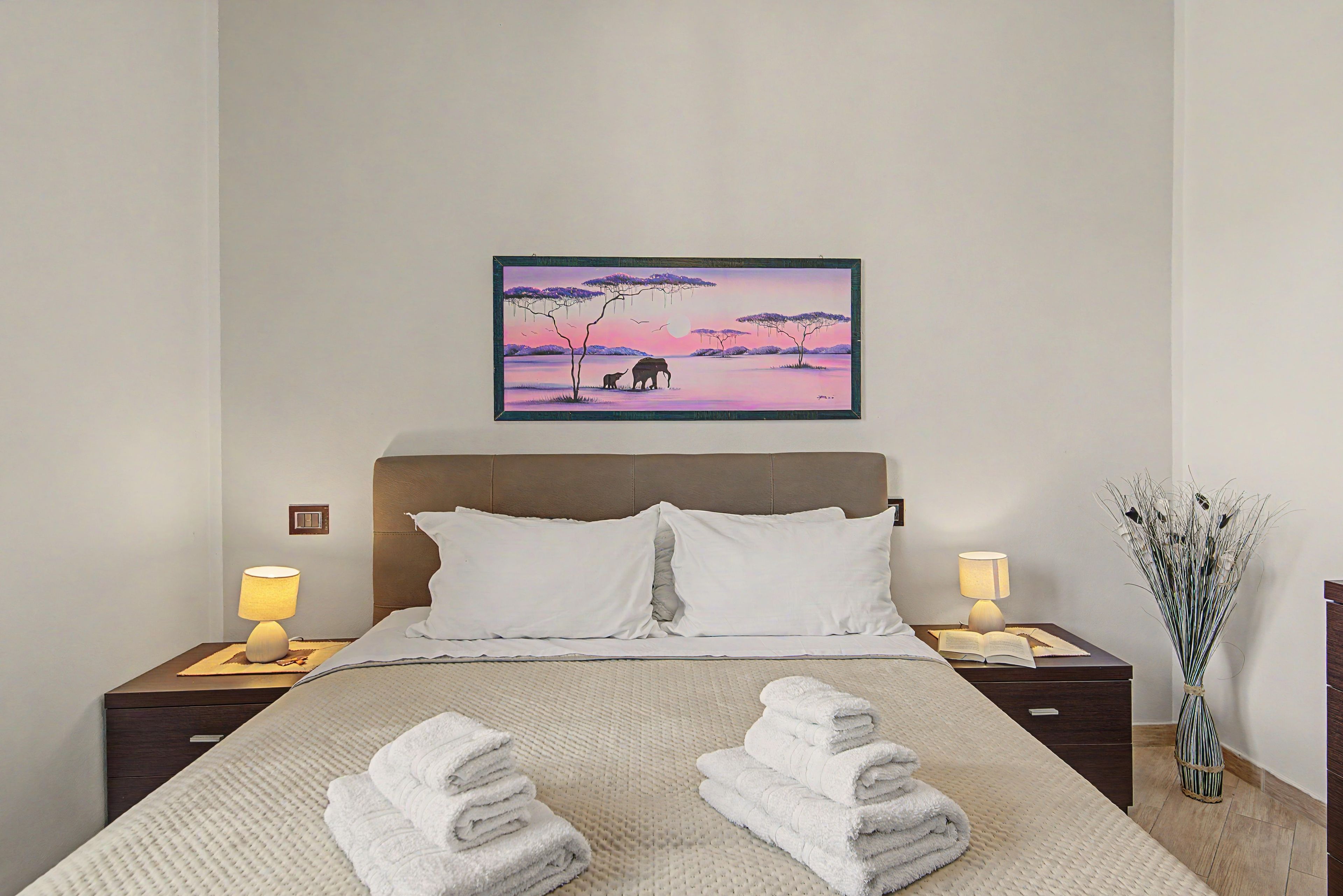 Una camera da letto con un letto, due lampade da comodino, un comodino con una lampada, un quadro appeso al muro e un vaso con piante.