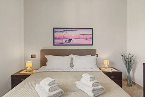 A bedroom with a bed, two bedside lamps, a nightstand with a lamp, a framed picture on the wall, and a vase with plants.
