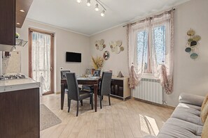 A modern living room with a dining area, a flat-screen TV mounted on the wall, and a large window with sheer curtains.