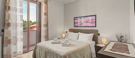 Habitación de hotel con cama, mesita de noche con lámpara, un cuadro en la pared y vistas al exterior.