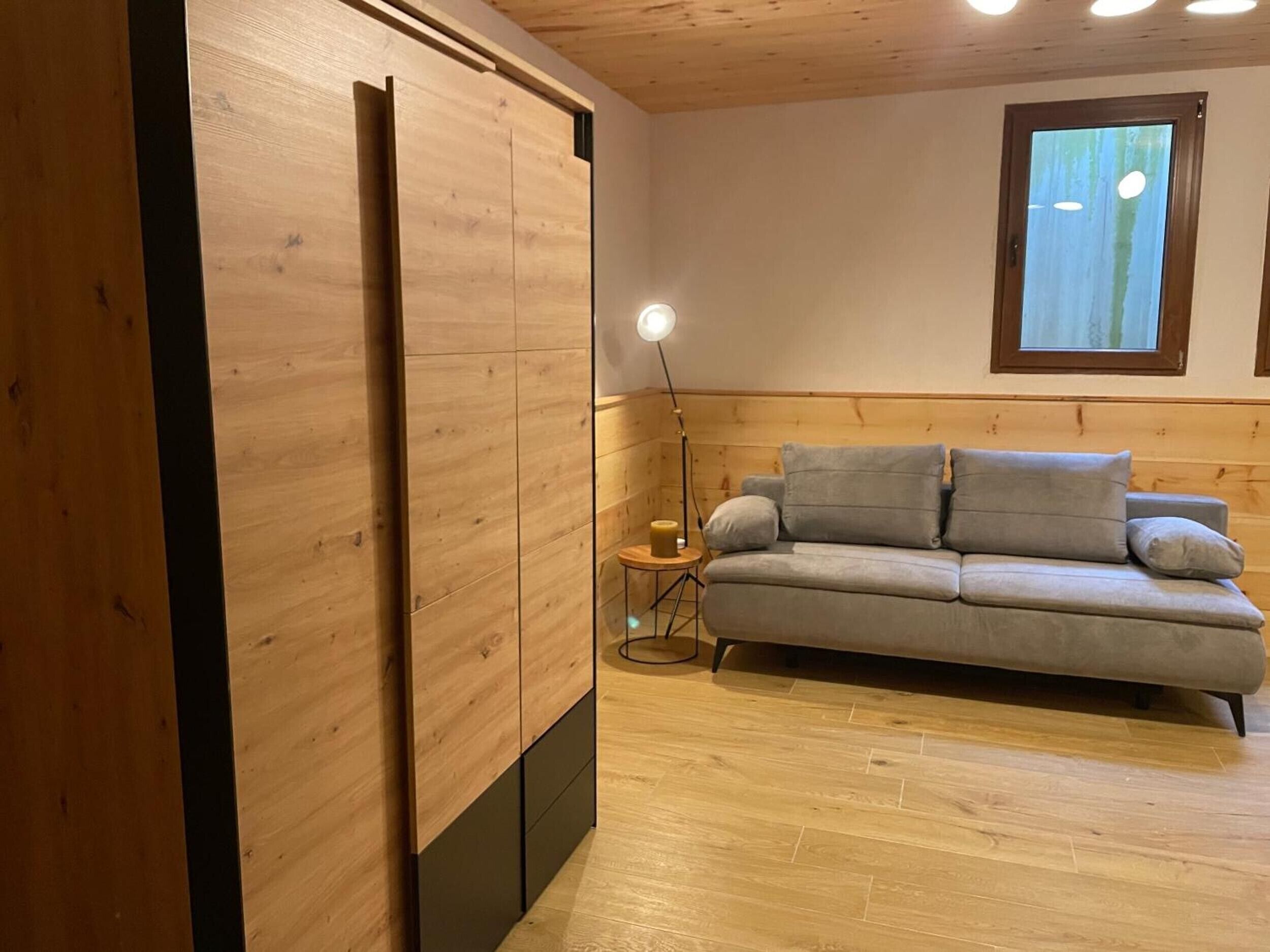A room with a wooden floor, a grey sofa, a small side table with a lamp, and a window with blinds.