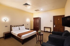 Ein Hotelzimmer mit Bett, Fernseher, Schrank, einem kleinen Tisch mit einer Vase und einem Sofa.
