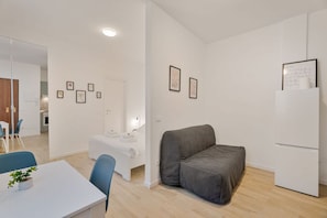 Ein modernes, minimalistisches Zimmer mit einem Bett, einem grauen Sofa, einem weißen Tisch mit Stühlen und einem weißen Kühlschrank.