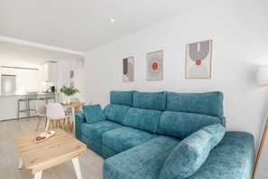 Ein modernes Wohnzimmer mit einem türkisfarbenen Sofa, einem Holztisch und einem Essbereich mit weißem Tisch und Stühlen.