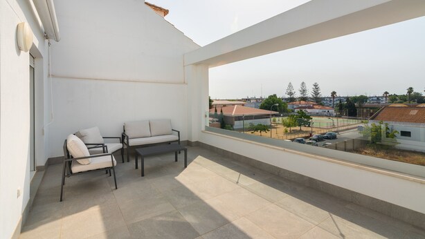 Un balcone con divano e poltrone, pavimento piastrellato e vista su una zona residenziale con case e alberi.