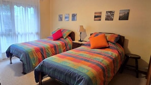 Ein Zimmer mit zwei Betten, jeweils mit bunter Bettwäsche, einem Nachttisch mit Lampe und einem kleinen Hocker.