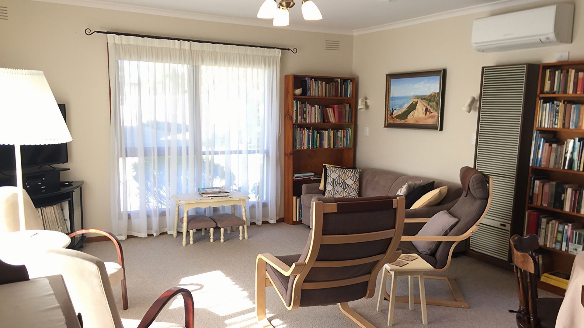A living room with a sofa, chairs, a table with books, a bookshelf, and a painting on the wall.