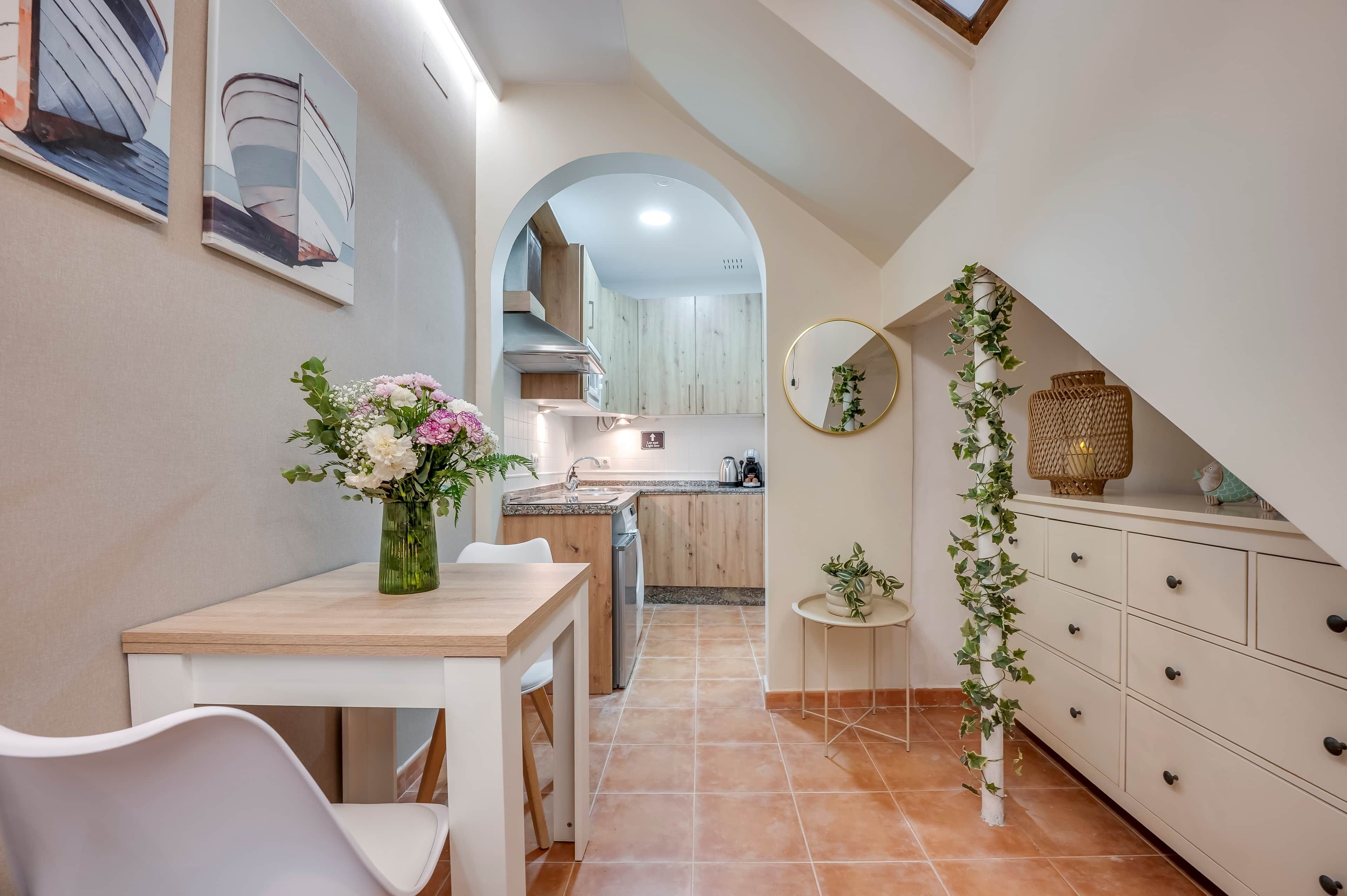 Una cocina estrecha con comedor, un armario blanco con cajones, un espejo redondo pequeño y un jarrón con flores sobre la mesa.