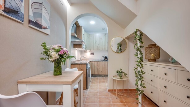 Una cocina estrecha con comedor, un armario blanco con cajones, un espejo redondo pequeño y un jarrón con flores sobre la mesa.