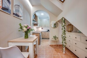 Eine schmale Küche mit Essbereich, einem weißen Schrank mit Schubladen, einem kleinen runden Spiegel und einem Blumenstrauß auf dem Tisch.