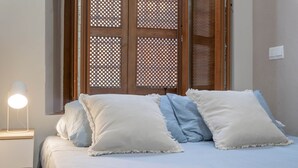 Una cama con almohadas blancas y azules, una lámpara de noche y un ventanal con un diseño de celosía de madera.
